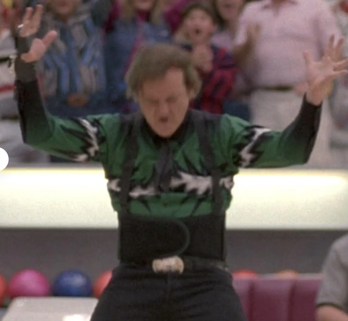 A man in a green shirt with a black belt and suspenders is celebrating with his arms raised in a bowling alley, surrounded by other people.