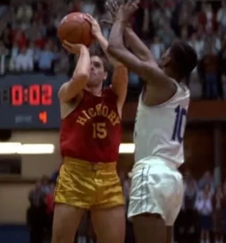 A basketball player wearing a red jersey with the number 15 is shooting a basketball, while an opposing player in a white jersey with the number 10 is attempting to block the shot.