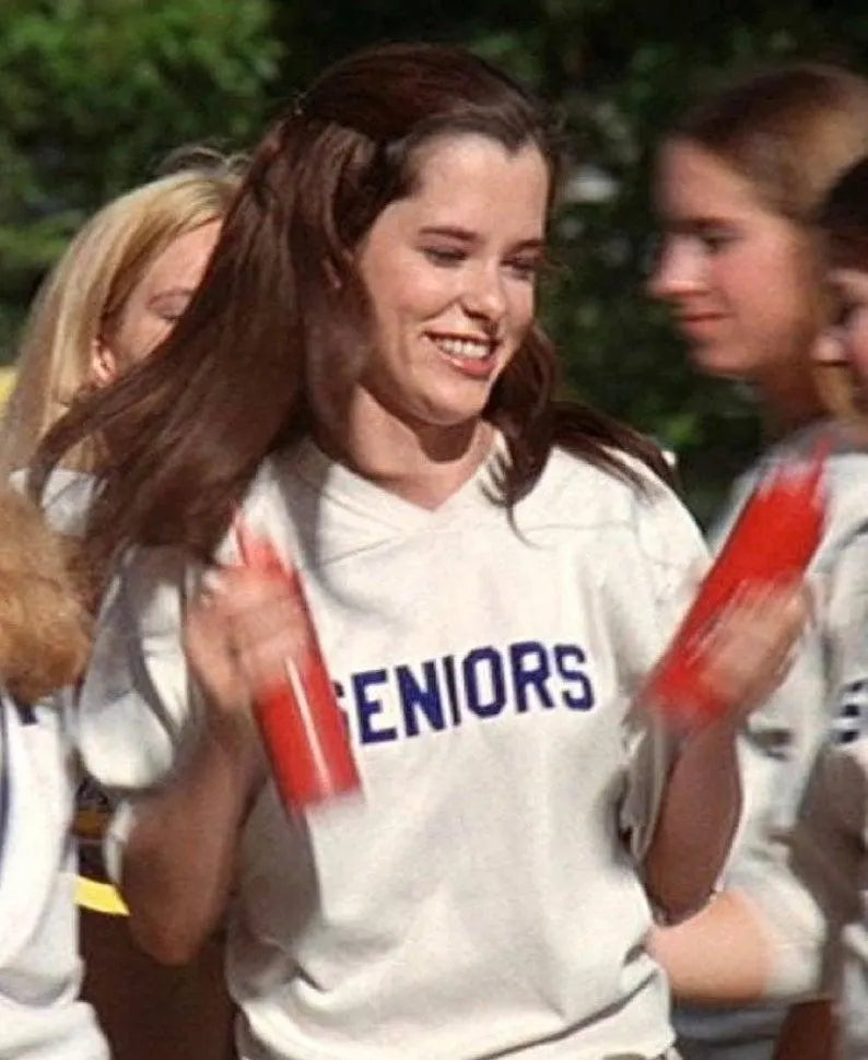 A young woman with long brown hair is smiling and holding a red object, possibly a water bottle or a sports accessory, while wearing a white shirt with the word "Seniors" written on it.