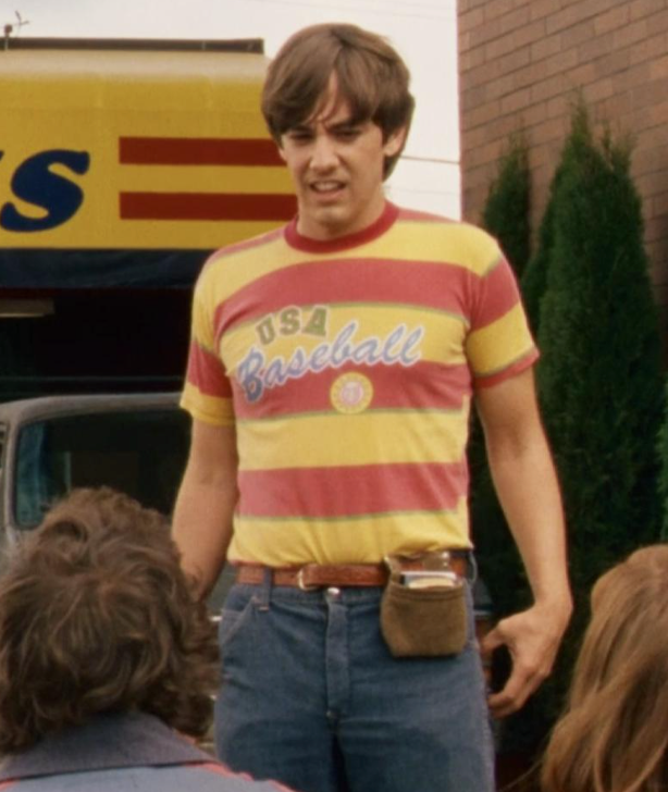 A man wearing a striped baseball shirt stands in front of a building, with a group of people behind him.