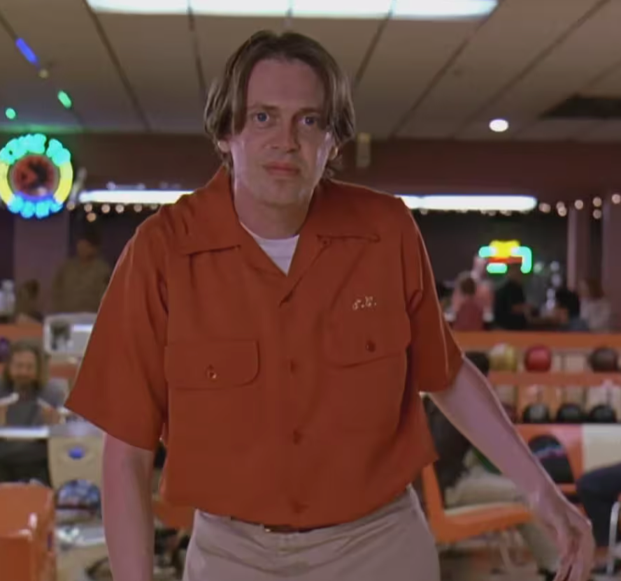 A man wearing a red shirt and khaki pants stands in a bowling alley, with a neon sign visible in the background.