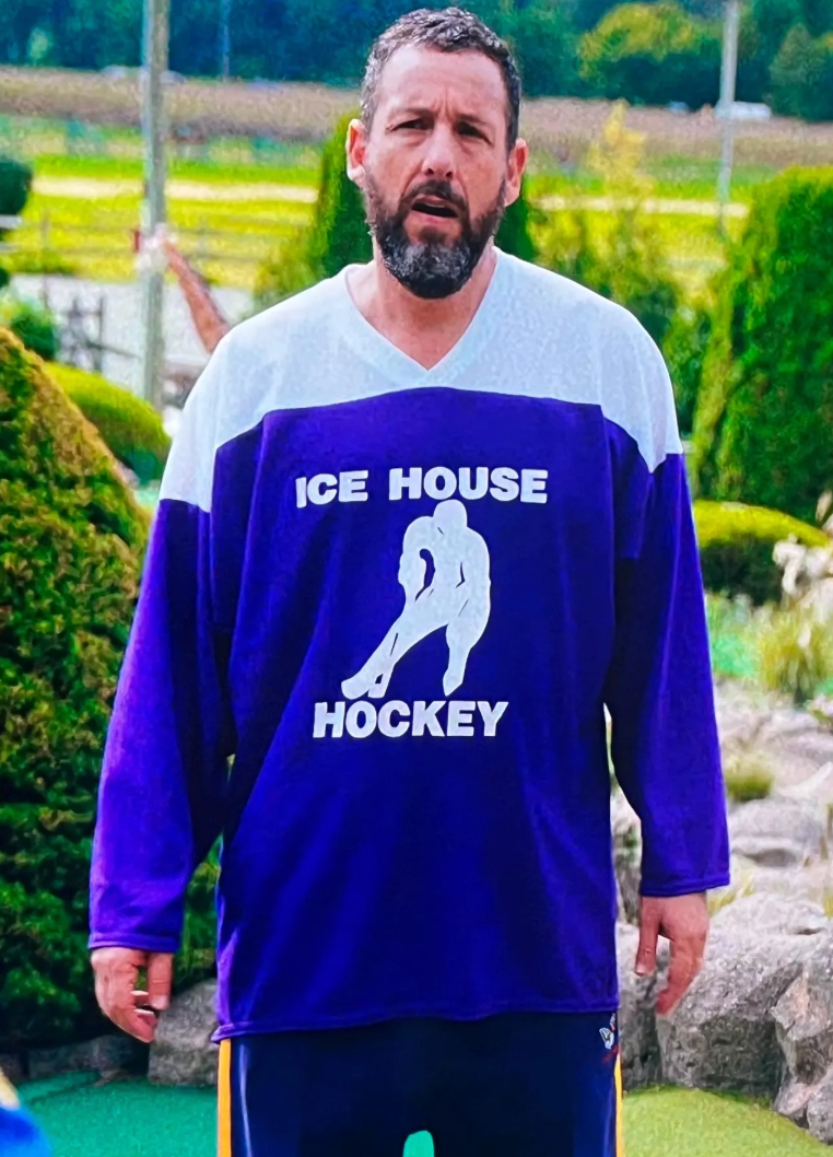 A man wearing a purple and white hockey jersey with the text "ICE HOUSE HOCKEY" printed on it, standing in a park-like setting with trees and bushes in the background.