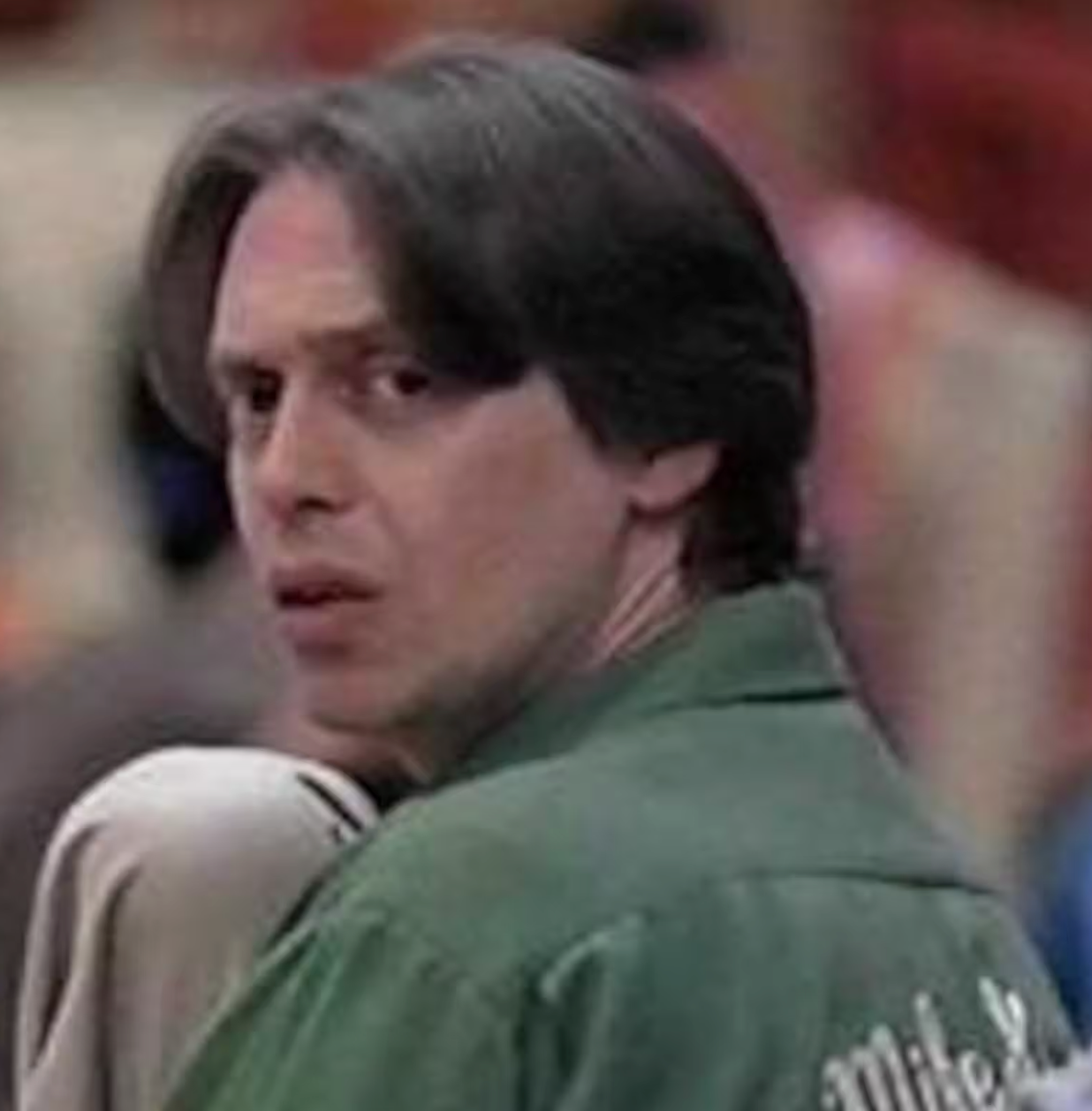 A man with short dark hair wearing a green shirt with white text on the back, looking off to the side with a serious expression.